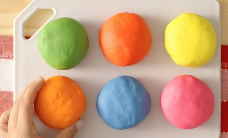 Seis bolas de massinha colorida em uma tábua branca, com uma mão pegando a laranja